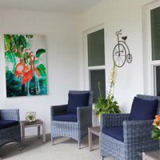 A cozy patio area featuring rattan chairs with navy cushions, vibrant artwork on the wall, decorative plants, and a small table for relaxation.