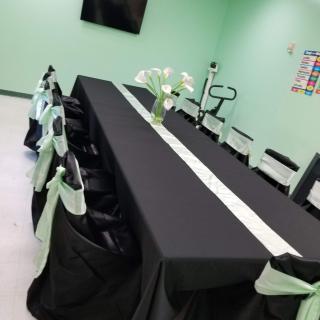 A conference room with a long meeting table covered in a black tablecloth and decorated with green accents. Chairs are neatly arranged with matching decor, and a vase of flowers sits at the center of the table.