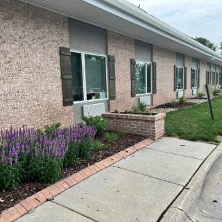 A brick building with multiple windows is surrounded by a landscaped area featuring flowers, bushes, and a sidewalk.