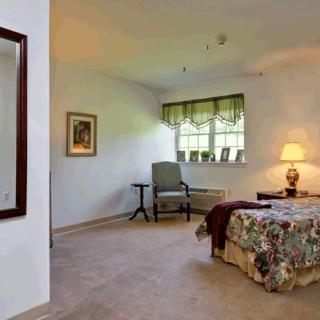 A cozy bedroom with a patterned bedspread, a chair by the window, and warm lighting from a table lamp.
