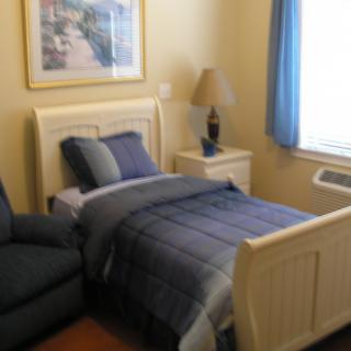 A cozy bedroom with a single bed featuring blue bedding, a nightstand with a lamp, and a window with blue curtains.