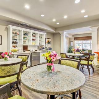 A modern dining area with round tables adorned with colorful flowers and a kitchenette in the background.