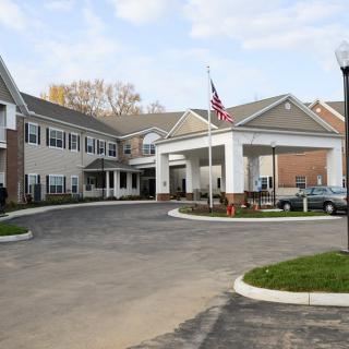 The image shows a large building with an entrance driveway and several parked cars, featuring a flagpole with an American flag.