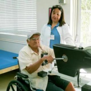 An elderly man in a wheelchair receives physical therapy assistance from a healthcare professional.