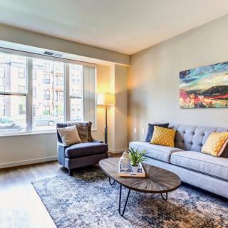 A cozy and modern living room with a sofa, armchair, and coffee table, complemented by colorful artwork and natural light streaming through the large window.