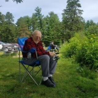 An elderly person sitting on a camping chair, fishing by a lakeside surrounded by lush greenery.