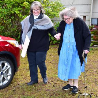 An elderly woman is assisted by another person as they walk together outside near a car.