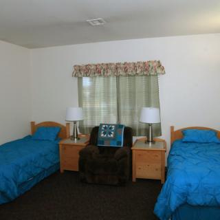 A simple bedroom with two twin beds and blue bedding, featuring a nightstand between the beds and a window with patterned curtains.