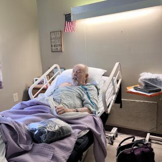 An elderly patient resting in a hospital room, surrounded by blankets and personal items.
