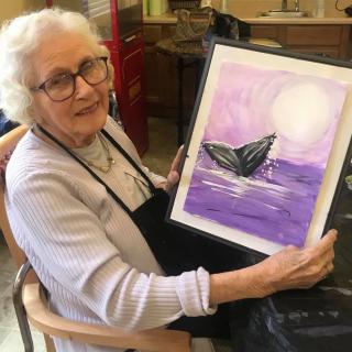 An elderly woman proudly displays her painting of a whale against a purple ocean backdrop.