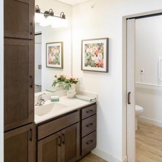 A modern bathroom with a wooden cabinet, artwork on the wall, and a vase of flowers on the counter.