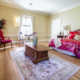 A cozy bedroom with red bedding, wooden floors, and natural light streaming in.