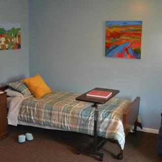 A simple bedroom with a single medical bed, colorful paintings on the wall, and slippers beside the bed.