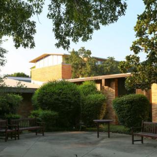 A peaceful outdoor area with benches and tables, surrounded by lush greenery and a modern building in the background.