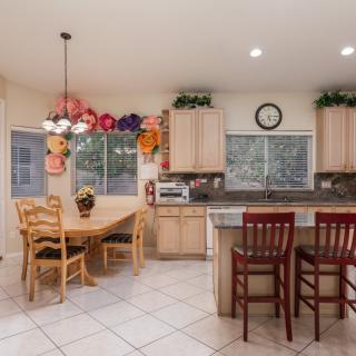 A cozy kitchen with a dining area, featuring a wooden table and chairs, a countertop with bar stools, and colorful decorative flowers.