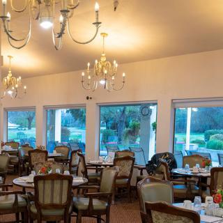 An elegant restaurant dining room featuring chandeliers, tables, and chairs with large windows overlooking a garden.