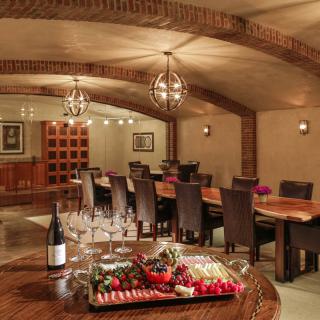 A luxurious dining area set in a wine cellar with a large table, elegantly arranged glasses, and a food platter.