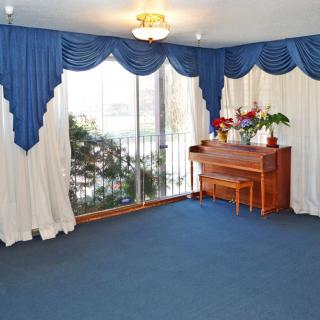 A living room featuring a piano, large windows with blue and white draped curtains, and a blue carpeted floor.