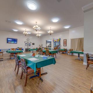 A spacious dining room with tables covered in green tablecloths, surrounded by chairs and illuminated by ceiling lights.