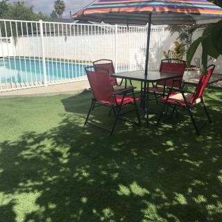 A sunny outdoor patio area with a table, chairs, and an umbrella near a swimming pool.