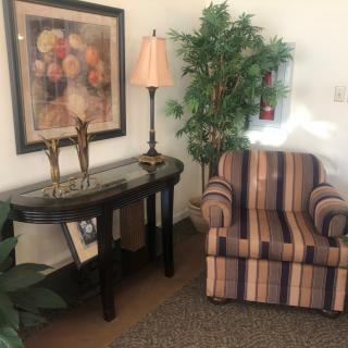 A cozy living room setup with a striped armchair, a sofa table adorned with a lamp and decorative items, artwork on the wall, and indoor plants.