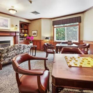 A cozy living room with leather chairs, a wooden table set for checkers, a fireplace, and natural light streaming through a large window.
