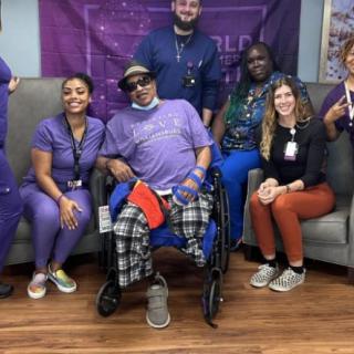A diverse group of healthcare workers pose for a cheerful group photo with a patient in a wheelchair.