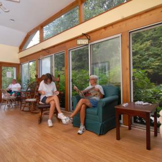 People relaxing and reading in a sunlit room with large windows.