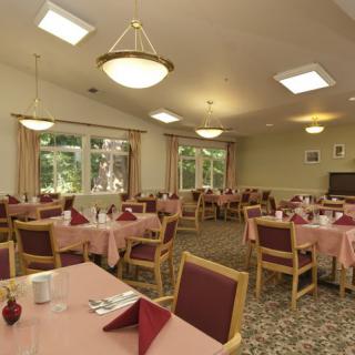 A spacious dining room with neatly arranged tables and chairs, adorned with pink tablecloths and illuminated by overhead lighting.