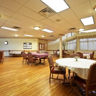 An empty dining room with wooden flooring, featuring arranged tables and chairs under bright ceiling lights.