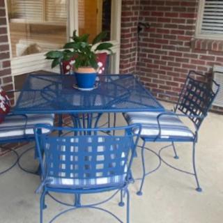 A cozy outdoor patio setup featuring a blue metal table and chairs with cushions, accompanied by a potted plant centerpiece against a brick backdrop.