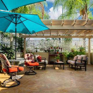 A cozy outdoor patio with comfortable seating and a vibrant umbrella under a pergola surrounded by lush greenery.