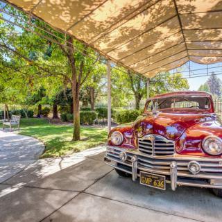 A classic red vintage car parked under a shade canopy on a sunny day, surrounded by lush greenery.
