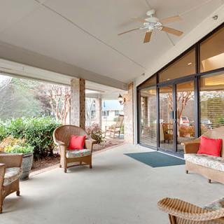 A cozy covered porch with wicker chairs and a ceiling fan, overlooking a garden through large glass doors.