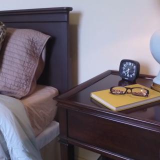 A cozy bedroom setup with a nightstand featuring a lamp, alarm clock, and reading glasses on top of a book.