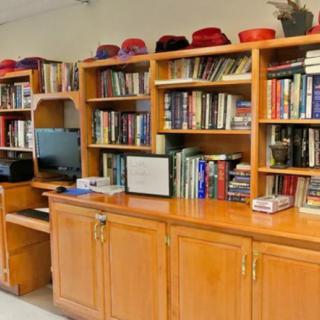 A wooden bookshelf filled with books and topped with a collection of hats, alongside a computer station.