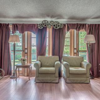 A cozy living room with two armchairs, flanked by elegant curtains and standing lamps, overlooks a lush garden through large windows.