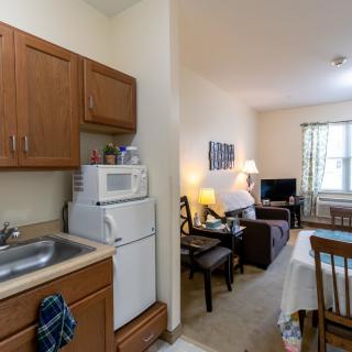 A cozy apartment with an open kitchen and living room layout, featuring wooden cabinets, a microwave, a dining table, and comfortable seating.