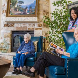 Two seniors enjoy a relaxing moment in a cozy living room, while a caregiver stands nearby.
