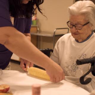 A caregiver assists an elderly woman with a cooking activity.
