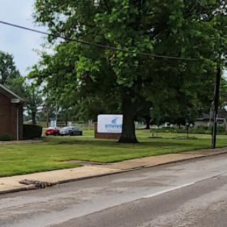 A view of a grassy area with a tree, a building on the left, and a sign in front.