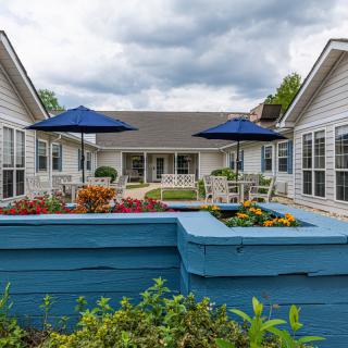 A residential courtyard featuring vibrant gardens with blue planters, outdoor seating, and umbrellas under a cloudy sky.
