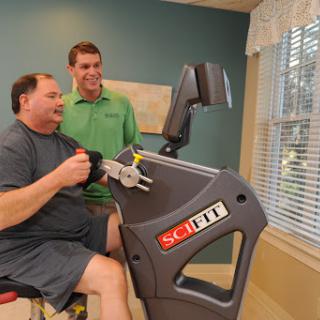 A man engages in a rehabilitation exercise with the assistance of a trainer, using specialized gym equipment.