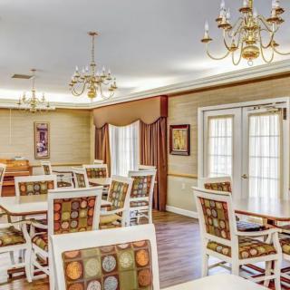 A well-lit dining room with elegant chandeliers, patterned chairs, and tables, featuring large windows with curtains and wood flooring.