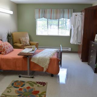 A cozy and well-lit hospital room designed for patient comfort, featuring an orange bedspread, a chair, and a window that brings in natural light.
