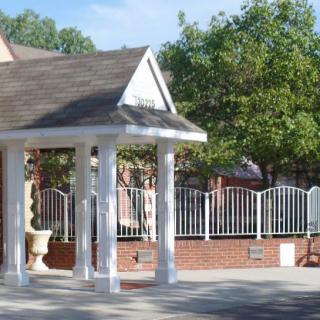 A small gazebo with white pillars stands near a brick wall and metal fence, surrounded by trees.