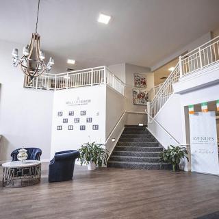 A spacious lobby interior featuring a central staircase, elegant chandelier, and seating area with plants.