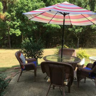 A cozy patio setup with a round table, cushioned chairs, and a colorful striped umbrella in a lush backyard.