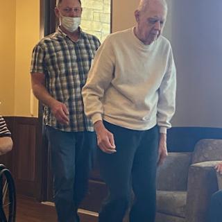 An elderly man assisted by a caregiver as they walk indoors.