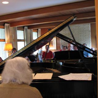 A group of people gathered around a grand piano inside a cozy room.